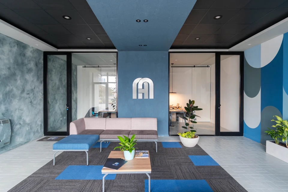 Recepción con un plano azul que cruza el cielorraso negro y baja sobre la pared, un monograma metálico en forma de arco, mamparas de aluminio y vidrio, y un mural de arcos en el fondo