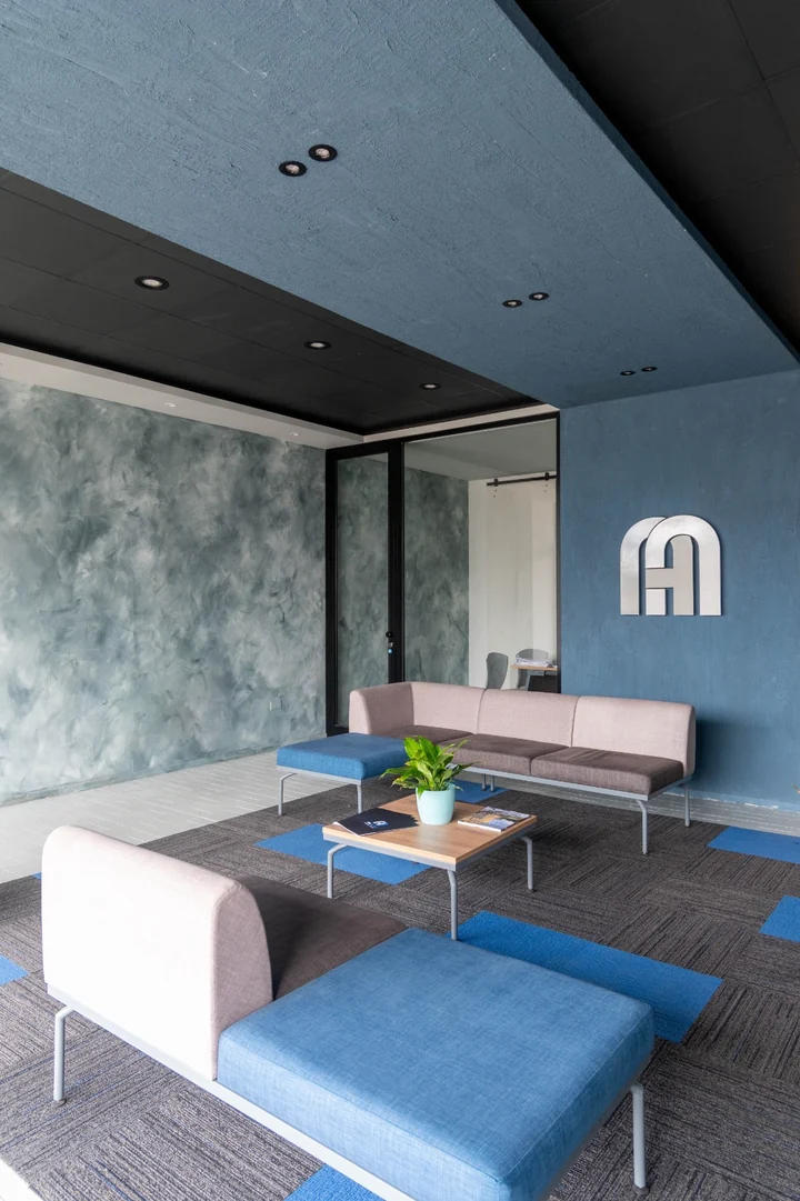 Sala de espera vista de costado, con el plano azul del cielorraso, la pared en veladura verde grisada y el monograma metálico