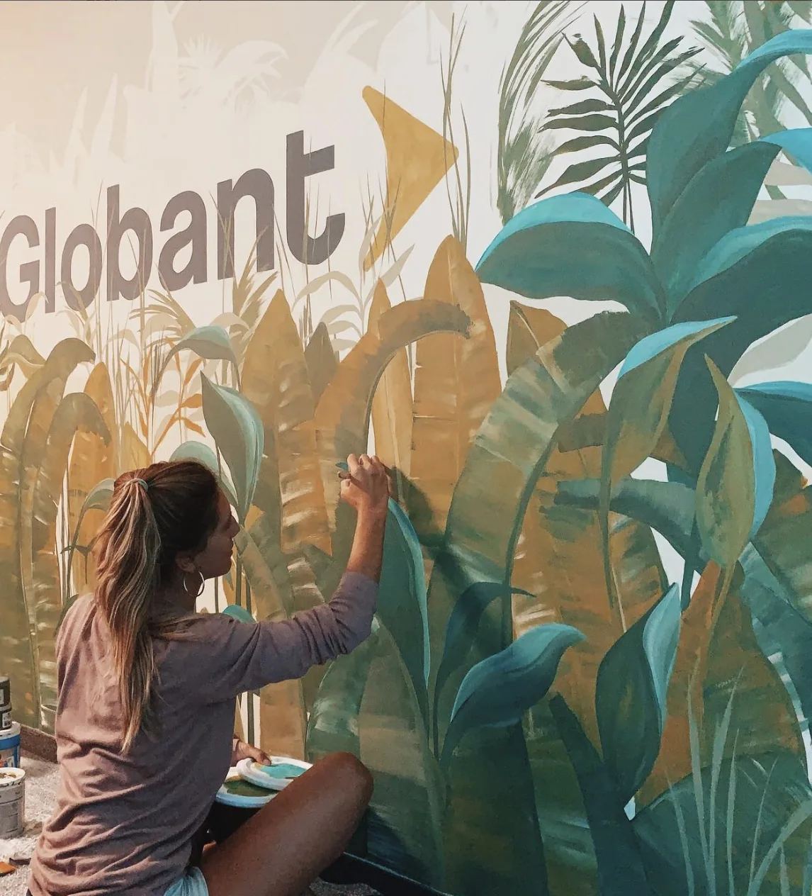 Belén Marcuzzi pintando un mural botánico en la pared de una oficina, con la marca Globant integrada al follaje