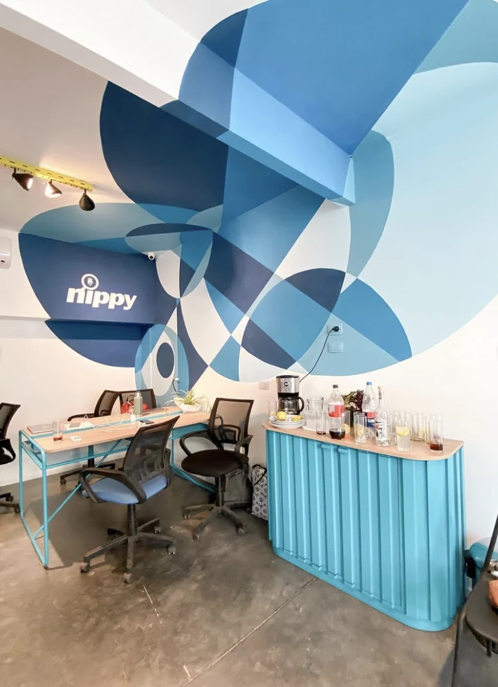 Interior de oficina con un gran mural geométrico de círculos superpuestos en azules, que recorre la pared, la esquina y la cara inferior de una viga, con el logotipo de Nippy pintado en la zona más oscura